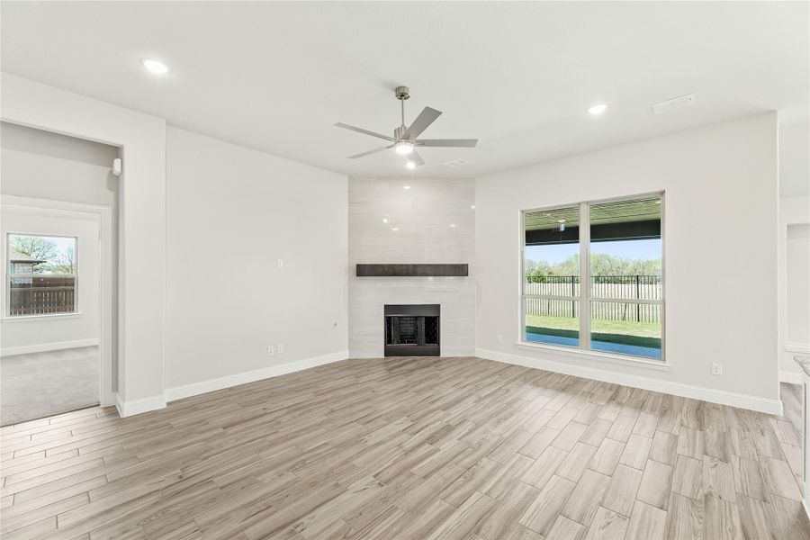 Open living room with light wood floors, ceiling fan, shiplap fireplace, and large backyard-facing window