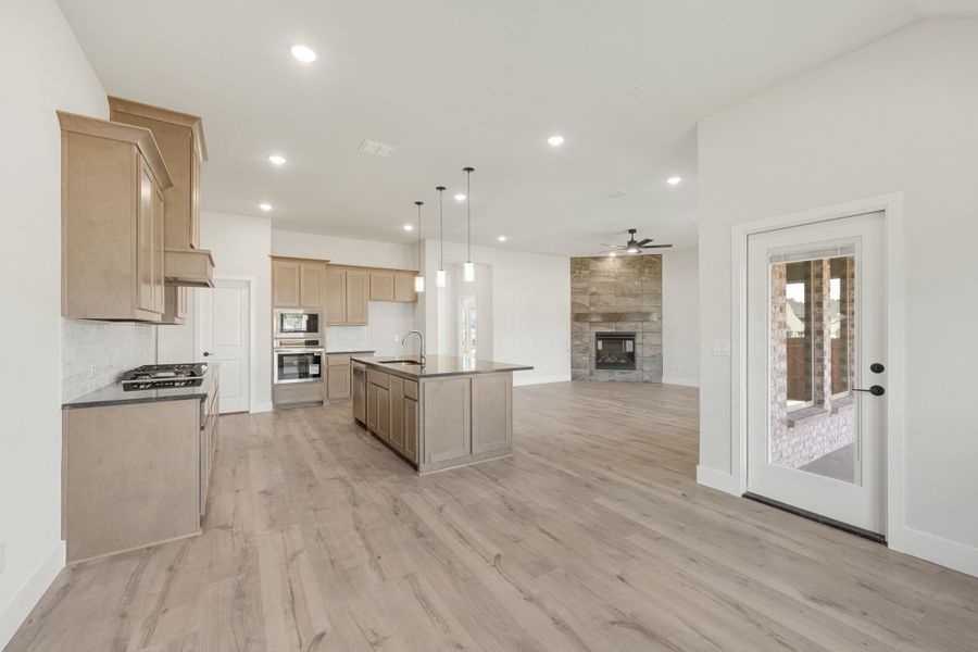 Open-concept kitchen with center island, tan cabinets, and light wood floors flowing into living area with stone fireplace.
