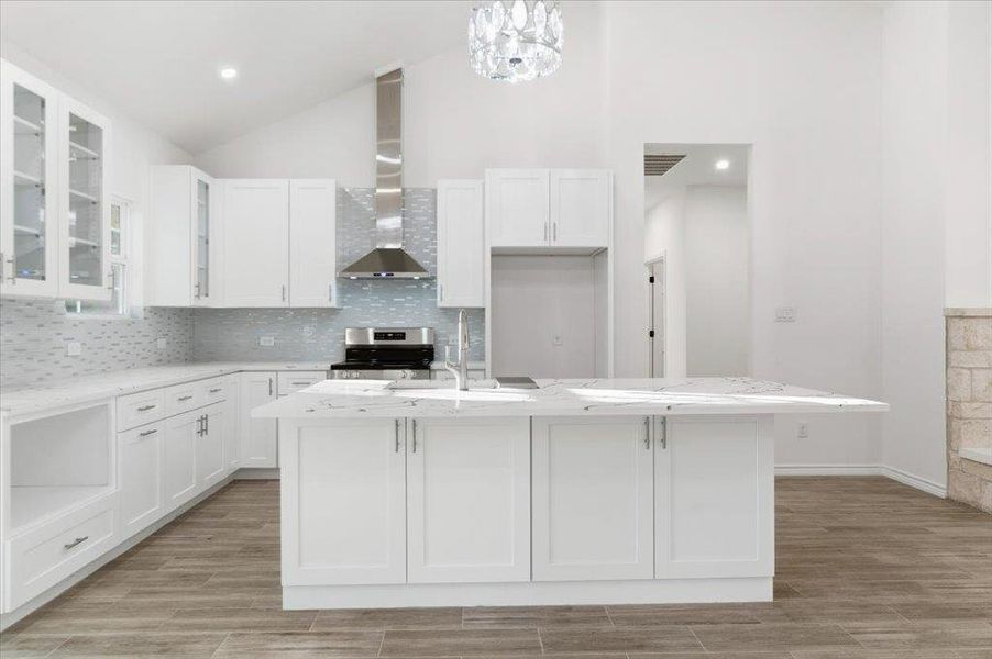 Kitchen with glass insert cabinets, light stone countertops, an island with sink, decorative backsplash, and stainless steel stove