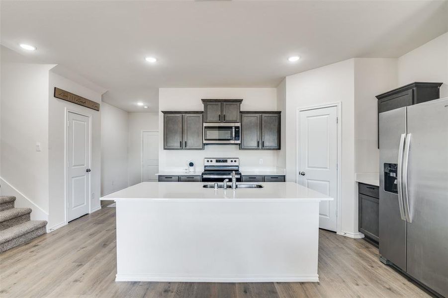 Kitchen featuring stainless steel appliances, an island with sink, light wood-style flooring, recessed lighting, and dark wood finish cabinetry Kitchen featuring stainless steel appliances, an island with sink, light wood-style flooring, recessed lighting, and dark wood finish cabinetry