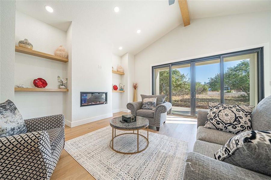 Living room with light wood-style floors, a glass covered fireplace, beam ceiling, recessed lighting, and high vaulted ceiling