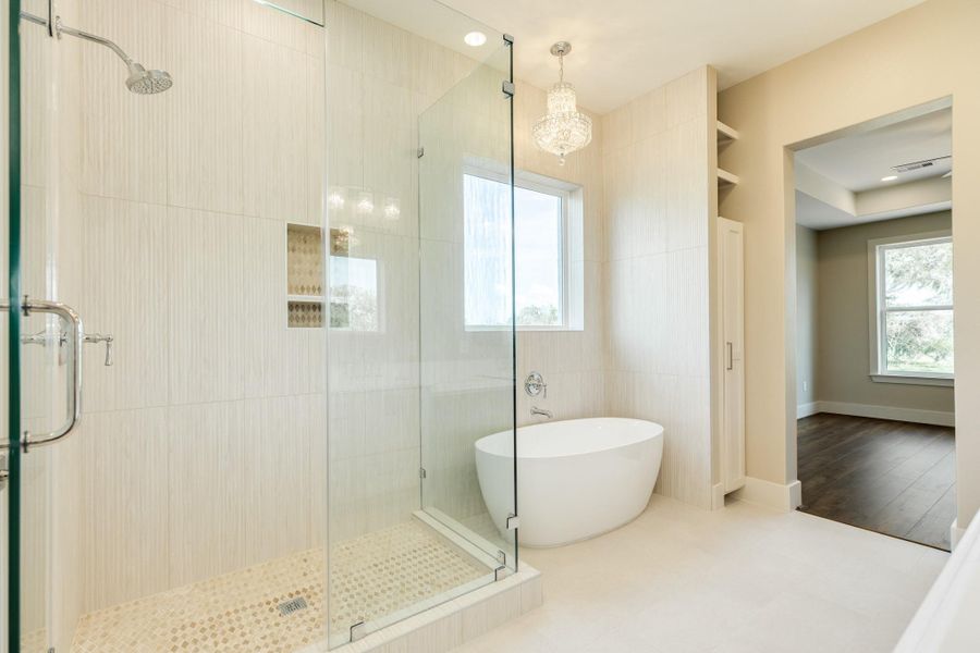 This photo showcases a modern bathroom featuring a spacious glass-enclosed shower and a freestanding bathtub. The room is well-lit with a large window and a chandelier, complemented by light-colored tiles and an adjacent area with hardwood flooring.
