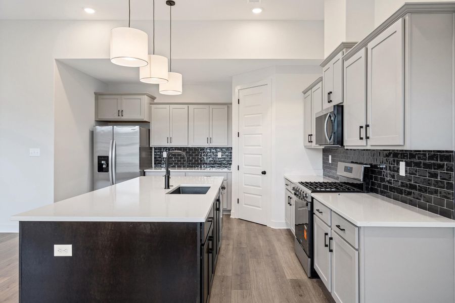 Kitchen featuring an expansive island with a built-in sink, white countertops, stainless steel appliances, light gray cabinetry, and a black subway tile backsplash Kitchen featuring an expansive island with a built-in sink, white countertops, stainless steel appliances, light gray cabinetry, and a black subway tile backsplash