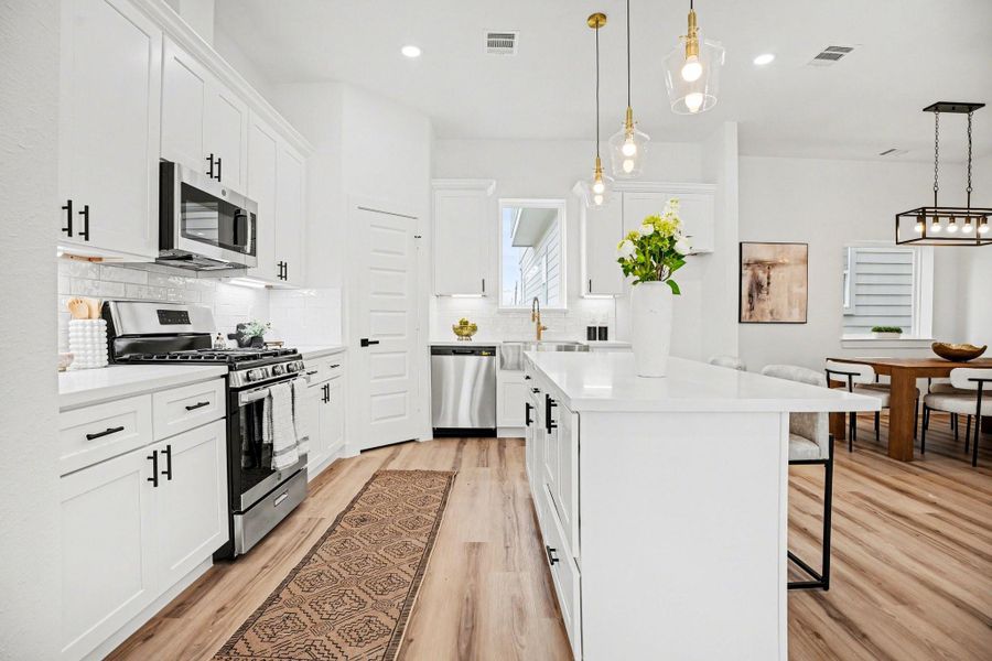 This kitchen features a modern design with white cabinetry, stainless steel appliances, and a central island with pendant lighting. The open layout seamlessly connects to a dining area with contemporary furnishings and stylish light fixtures, creating an inviting and functional space.