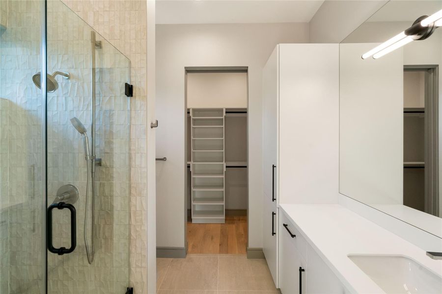 This modern bathroom features a sleek glass-enclosed shower with textured tiles, a spacious vanity with clean lines, and ample storage. It leads to a walk-in closet with organized shelving, enhancing functionality and style.