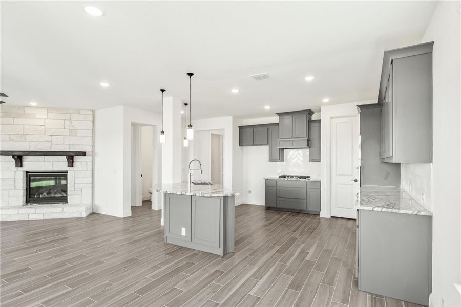 Open kitchen with gray cabinets, center island, marble countertops, and wood-look tile floors adjacent to a stone fireplace. Open kitchen with gray cabinets, center island, marble countertops, and wood-look tile floors adjacent to a stone fireplace.