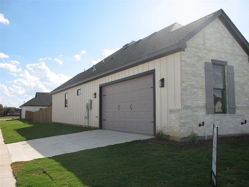 View of side of home featuring board and batten siding, driveway, brick siding, an attached garage, and a yard