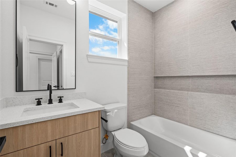 The third full bathroom features a warm wood vanity, a white quartz countertop, and a shower-tub combination. A large vertical window brings in excellent natural light, complemented by a durable, textured tile surround.