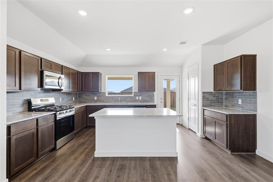 Kitchen featuring stainless steel appliances, light stone counters, dark brown cabinetry, lofted ceiling, and a kitchen island Kitchen featuring stainless steel appliances, light stone counters, dark brown cabinetry, lofted ceiling, and a kitchen island