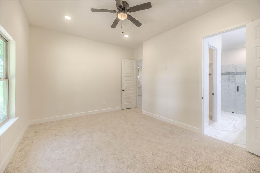 Another view of the primary bedroom. Bright and spacious bedroom with plush carpet, ceiling fan, large window, and an en-suite bathroom