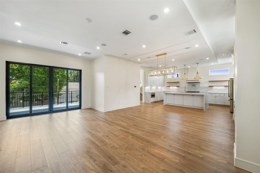Open-concept living, dining and kitchen area with wood floors and modern lighting. Large sliding glass doors lead to an outdoor balcony, offering plenty of natural light. The kitchen features sleek white cabinetry, a large island, and contemporary pendant lights, creating a stylish and functional space.