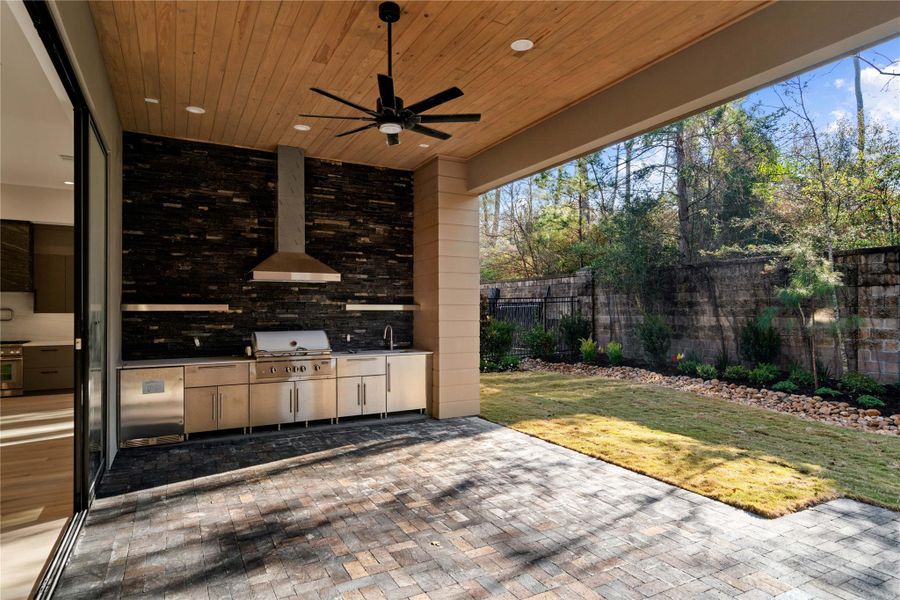 This photo showcases a modern outdoor kitchen and patio area. The space features a built-in grill, stainless steel cabinetry, and a sleek black backsplash. The ceiling is wood-paneled with recessed lighting and a ceiling fan. It opens up to a landscaped backyard with a stone border, creating an inviting space for outdoor entertaining.