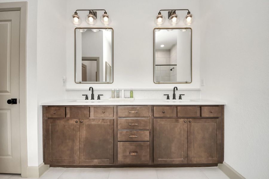 Spacious primary bathroom vanity featuring dual sinks, rich wood cabinetry, black fixtures, framed mirrors, and modern vanity lighting. The long countertop offers plenty of space for everyday routines, while the clean neutral finishes create a bright and polished feel.