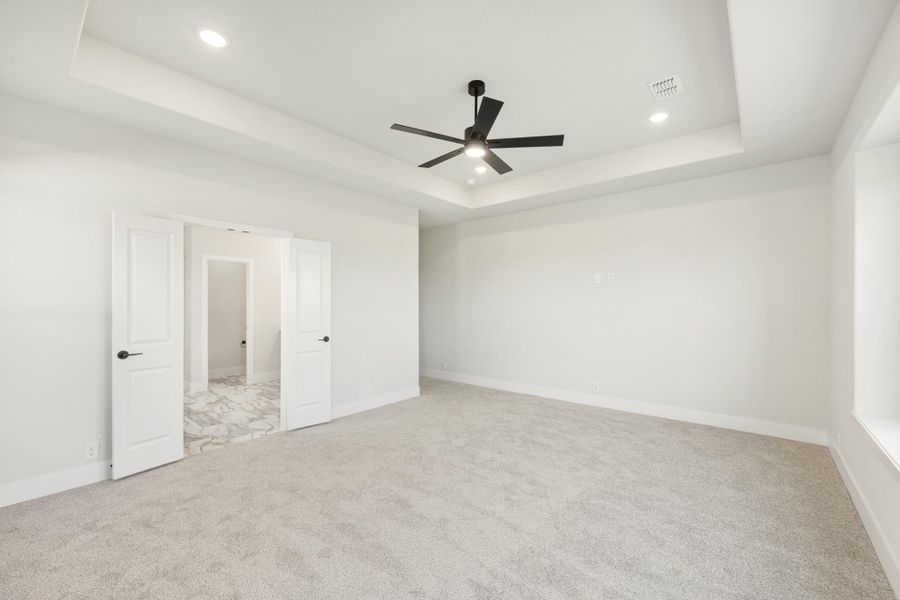 Empty bedroom with tray ceiling, black ceiling fan, carpet flooring, and open doors to an ensuite bathroom