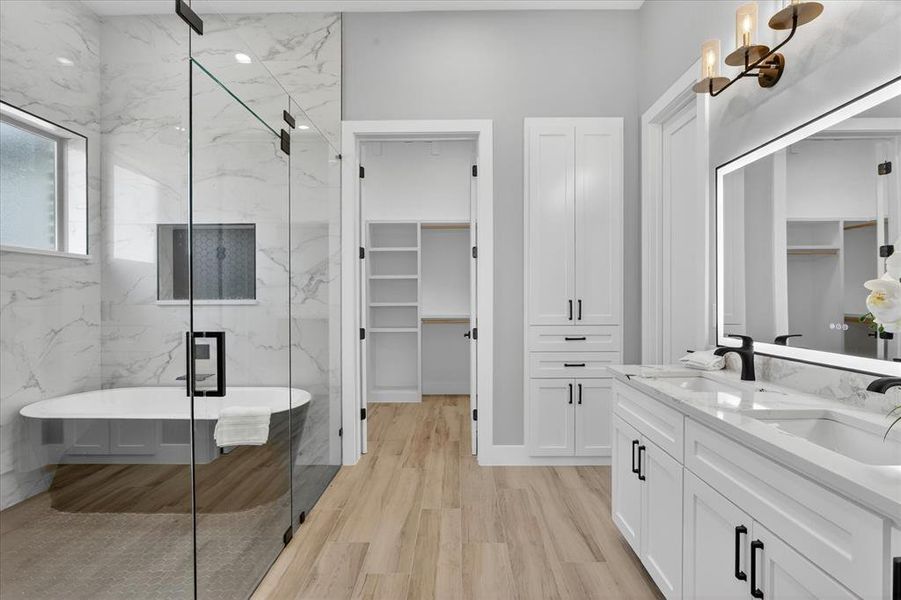 Bathroom featuring a dual vanity with quartz countertop, light wood-finish flooring, a freestanding soaking tub, glass-enclosed shower, and built-in white cabinetry