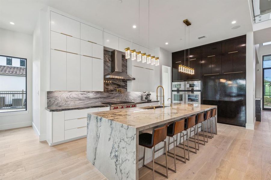 Kitchen with wall chimney range hood, decorative backsplash, a center island with sink, and light wood-type flooring