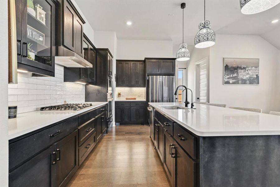 Kitchen featuring decorative light fixtures, tasteful backsplash, light wood-style floors, a center island with sink, and light stone counters Kitchen featuring decorative light fixtures, tasteful backsplash, light wood-style floors, a center island with sink, and light stone counters