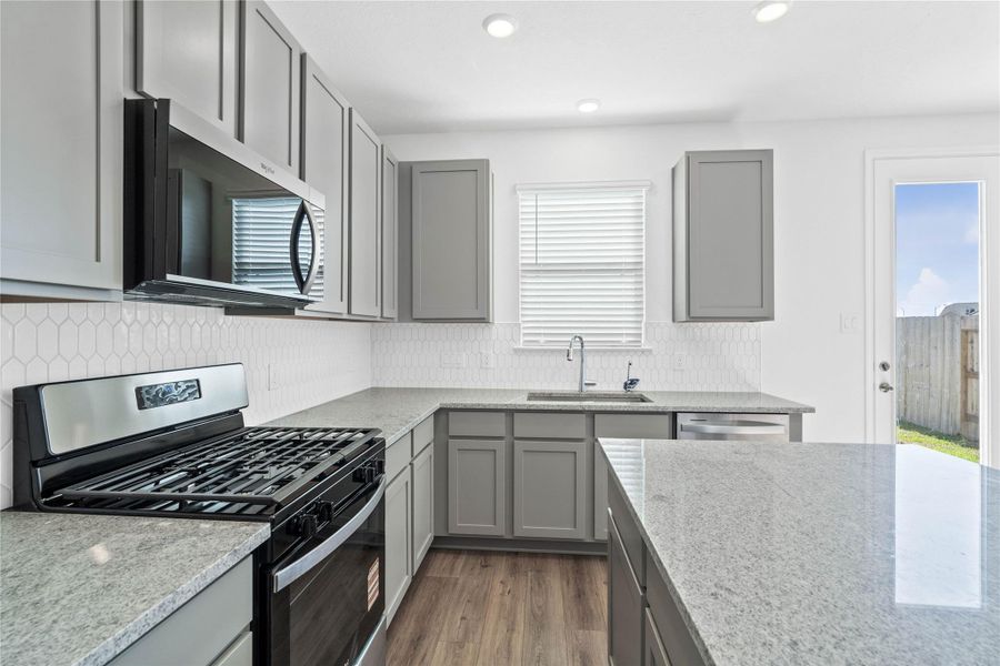 A true chef’s dream, this kitchen boasts high ceilings, recessed lighting, Woodmont Dakota cabinets in Pebble stain, Granite Summit White countertops, Whirlpool stainless steel appliances, a modern Daltile backsplash, and island—all with a clear view of the spacious nook.