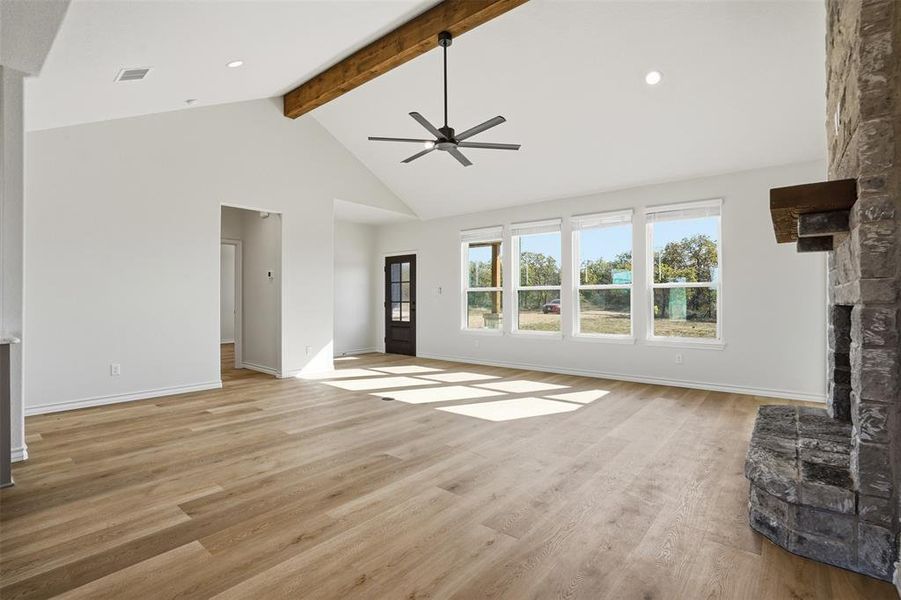Unfurnished living room with a stone fireplace, light wood-style floors, high vaulted ceiling, a ceiling fan, and beam ceiling Unfurnished living room with a stone fireplace, light wood-style floors, high vaulted ceiling, a ceiling fan, and beam ceiling