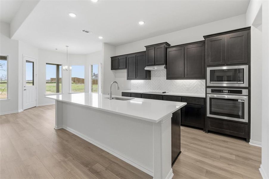 Kitchen with a kitchen island with sink, stainless steel appliances, light wood-style floors, suspended lighting, and decorative backsplash