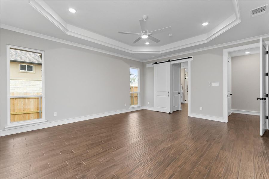 Unfurnished bedroom with a tray ceiling, a barn door, dark wood-style flooring, crown molding, and baseboards Unfurnished bedroom with a tray ceiling, a barn door, dark wood-style flooring, crown molding, and baseboards
