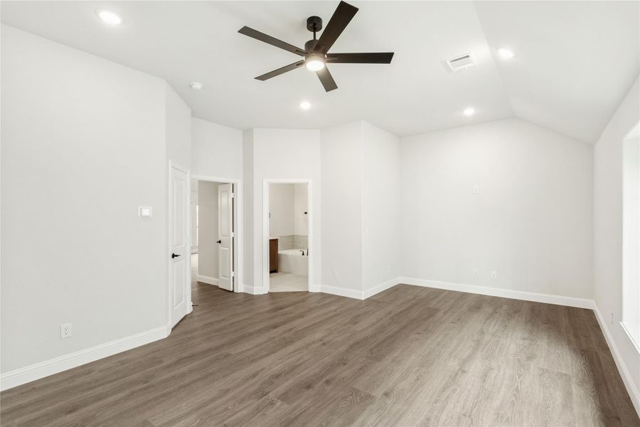 Empty master bedroom with vaulted ceiling, ceiling fan, wood floors, and ensuite bathroom doorway Empty master bedroom with vaulted ceiling, ceiling fan, wood floors, and ensuite bathroom doorway
