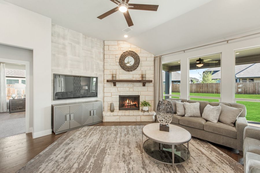 Living room with stone fireplace, vaulted ceiling, ceiling fan, and large windows overlooking backyard