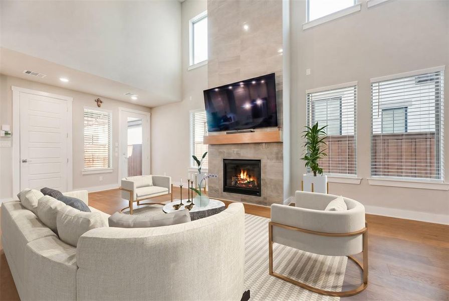 Two-story living area featuring a tile-surround fireplace with a wood mantle, wood-finish flooring, recessed lighting, and multiple windows with blinds