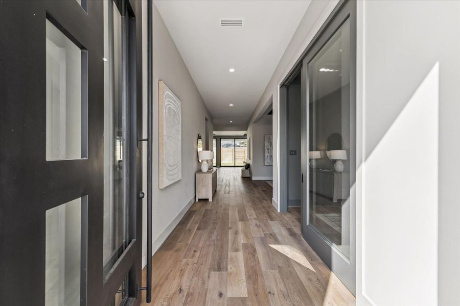 The entrance of the home makes a striking first impression with its modern glass and metal front door opening to a wide, light-filled hallway. Warm oak flooring extends the length of the space, drawing the eye toward the expansive living areas and backyard beyond, while sleek glass doors along the hall create a sense of openness and contemporary elegance.
