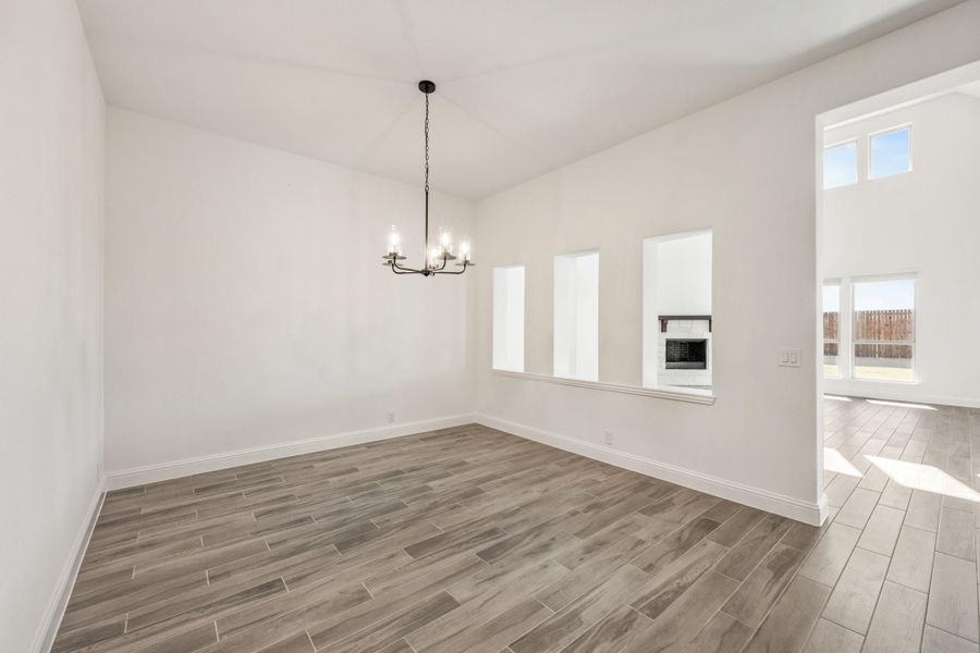 Empty dining room with wood-look tile floors, black chandelier, and white walls open to living area with fireplace Empty dining room with wood-look tile floors, black chandelier, and white walls open to living area with fireplace