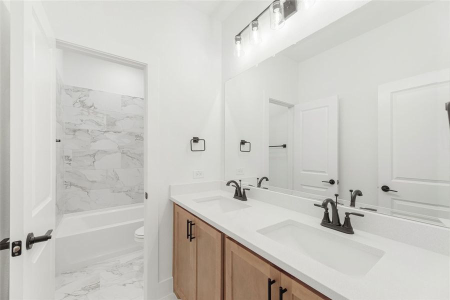 Bathroom with double sink vanity, wood cabinets, white countertop, and marble-tile tub surround with black fixtures Bathroom with double sink vanity, wood cabinets, white countertop, and marble-tile tub surround with black fixtures