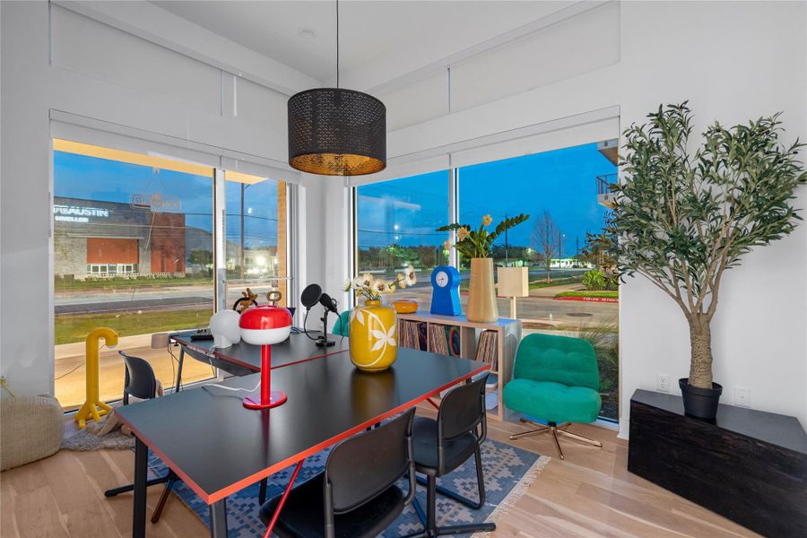 Expansive full-height windows illuminate a space featuring light wood-finish flooring, a modern black drum pendant light, and a built-in shelving unit