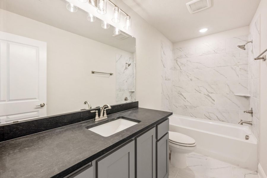 Bathroom with dark granite vanity, gray cabinets, and tub-shower combo with marble-style tile surround Bathroom with dark granite vanity, gray cabinets, and tub-shower combo with marble-style tile surround