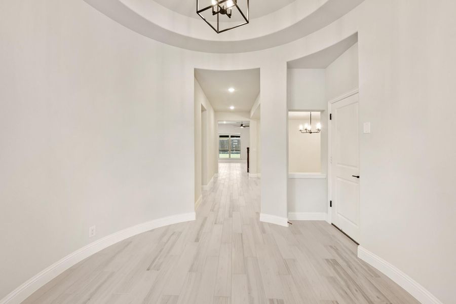Circular foyer with tray ceiling, black chandelier, and light hardwood floors leading to hallway Circular foyer with tray ceiling, black chandelier, and light hardwood floors leading to hallway