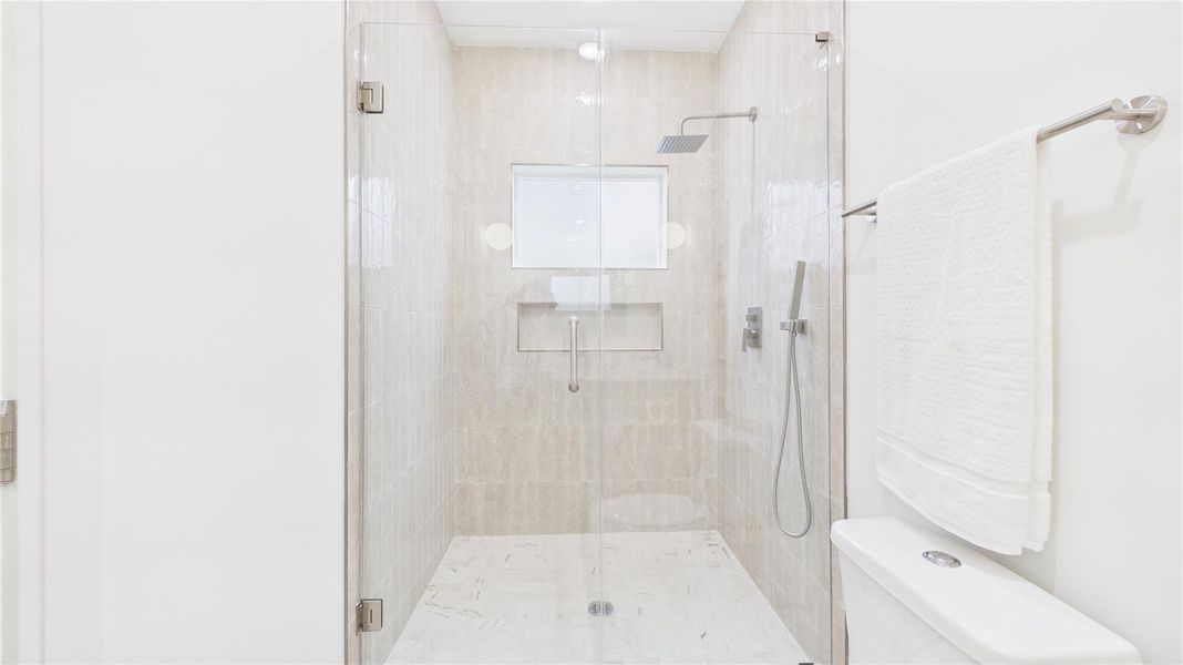 This is a modern, sleek bathroom with a walk-in glass shower featuring a rainfall showerhead. It has light beige tiles, a built-in shelf, and a clean, minimalist design. The white walls and fixtures add to the bright, spacious feel.