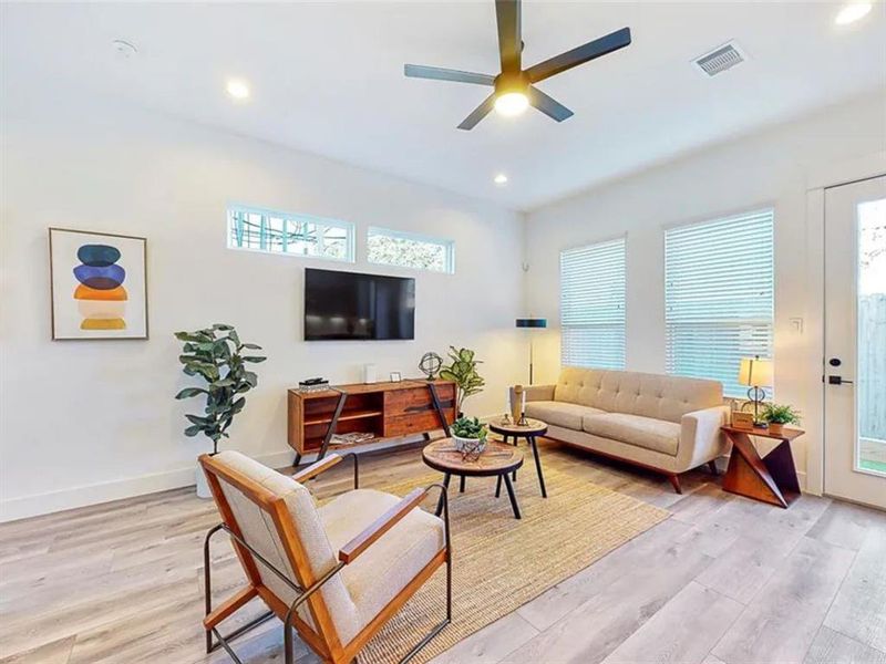 A modern and spacious living area with an open floor plan that seamlessly connects to the kitchen. Natural light floods the room, illuminating the warm wood floors and the contemporary furniture.