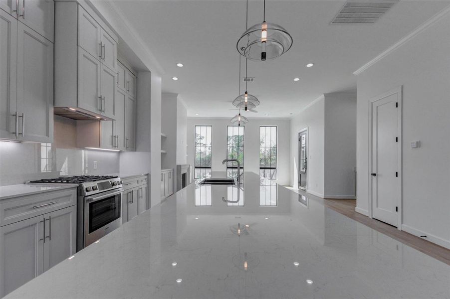 Striking oversized island with Dekton Malibu surface reflecting designer pendant lighting above. A deep single-basin sink and sleek faucet anchor the workspace while preserving clear views toward the tall black-framed windows. The open-concept design seamlessly connects kitchen, dining, and living areas, creating a bright and sophisticated entertaining space at the heart of this gated Memorial residence.