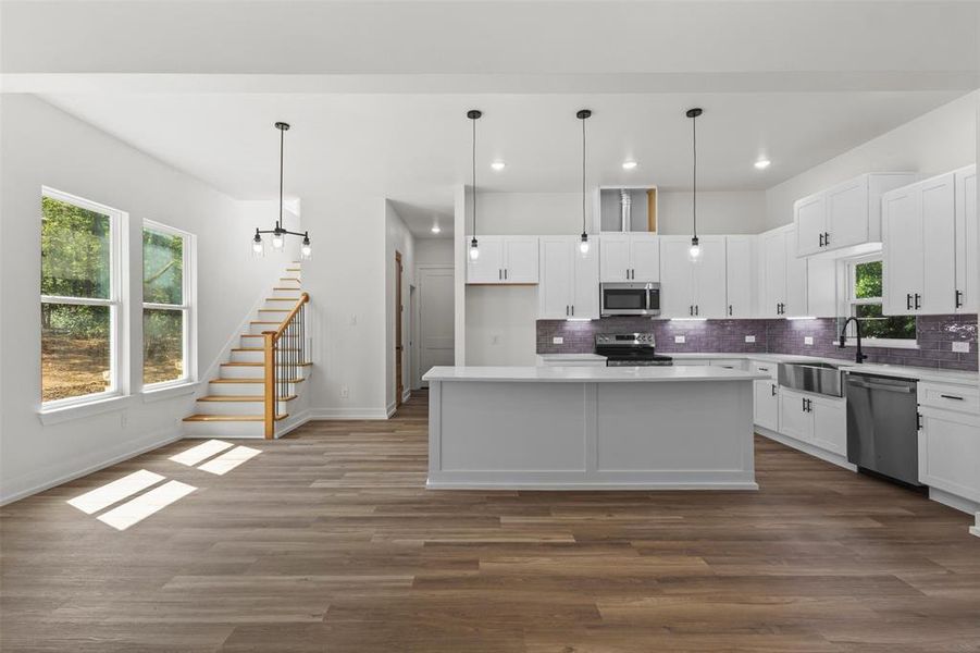 Open-concept living space featuring wide plank flooring, a modern white kitchen with stainless steel appliances, and a staircase with natural wood treads