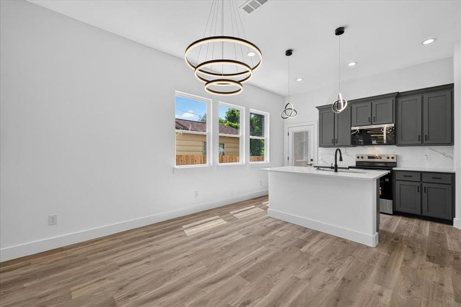 Open-concept living space featuring wood-finish flooring, a contemporary multi-ring chandelier, and a kitchen with dark cabinetry Open-concept living space featuring wood-finish flooring, a contemporary multi-ring chandelier, and a kitchen with dark cabinetry