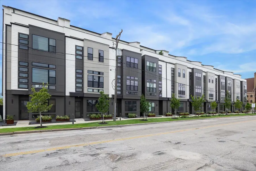 Modern townhouse complex featuring sleek, contemporary architecture with large windows and a mix of dark and light paneling. The street view shows well-maintained landscaping with young trees and a neatly trimmed lawn. Ideal for those seeking urban living with a stylish design.