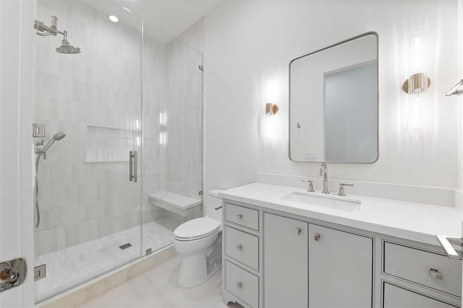 This is a modern, bright bathroom featuring a glass-enclosed walk-in shower, a sleek vanity with ample storage, and sophisticated gold-tone fixtures. The room is finished with large, light-tinted tiles that contribute to a clean, airy feel.