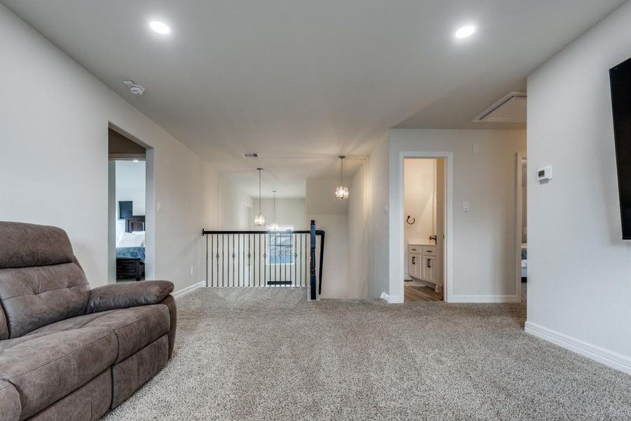 A cozy upstairs area/ Gameroom with plush carpet, featuring a seating nook and multiple doorways leading to bedrooms and a bathroom. The space is illuminated by recessed lighting and a hanging light fixture, with a railing overlooking the staircase.