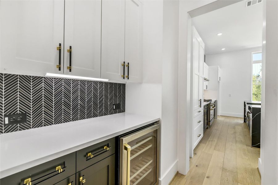 A sophisticated butler’s pantry featuring a striking herringbone backsplash, under-cabinet lighting, quartz counters, and custom two-tone cabinetry with matte black and brushed brass hardware. Includes a built-in wine fridge and seamless flow into the chef’s kitchen—perfect for entertaining in style. A sophisticated butler’s pantry featuring a striking herringbone backsplash, under-cabinet lighting, quartz counters, and custom two-tone cabinetry with matte black and brushed brass hardware. Includes a built-in wine fridge and seamless flow into the chef’s kitchen—perfect for entertaining in style.