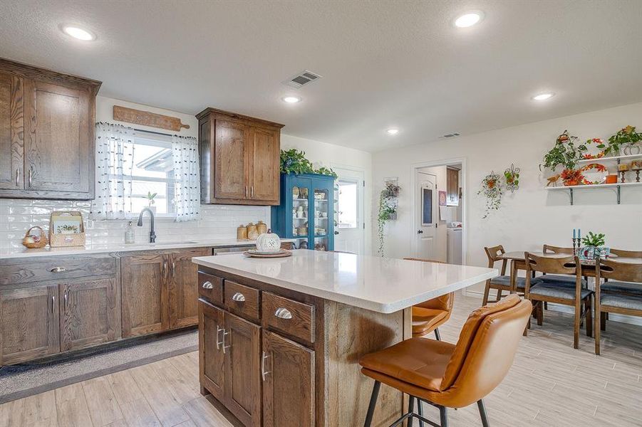 Kitchen featuring backsplash, a breakfast bar area, light wood-style tile flooring, a center island, and light quartz countertops Kitchen featuring backsplash, a breakfast bar area, light wood-style tile flooring, a center island, and light quartz countertops