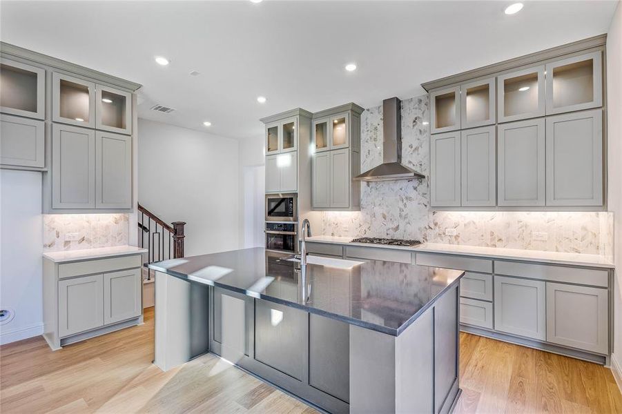 Kitchen featuring wall chimney range hood, a kitchen island with sink, backsplash, stainless steel appliances, and light wood-type flooring