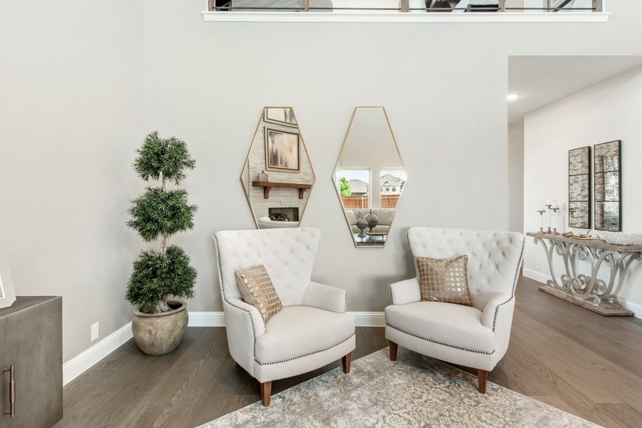 Sitting area with two white tufted accent chairs, geometric mirrors, and a potted topiary on hardwood floors