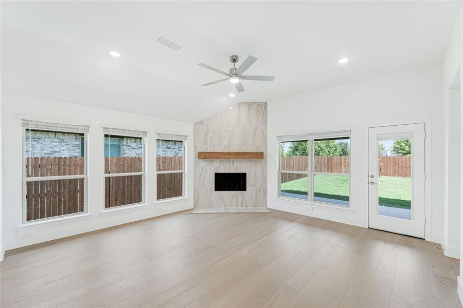 Open living room with light wood floors, marble tile fireplace, ceiling fan, and large windows