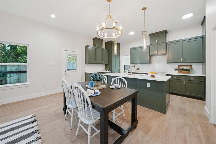 Kitchen featuring green cabinetry, decorative backsplash, light wood-style flooring, hanging light fixtures, and a kitchen island with sink