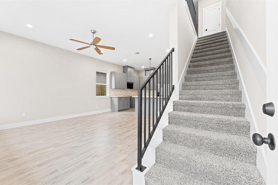 Spacious open-concept living area with light wood flooring, modern kitchen featuring grey cabinets and stainless steel appliances, and a cozy carpeted staircase leading to the upper level.