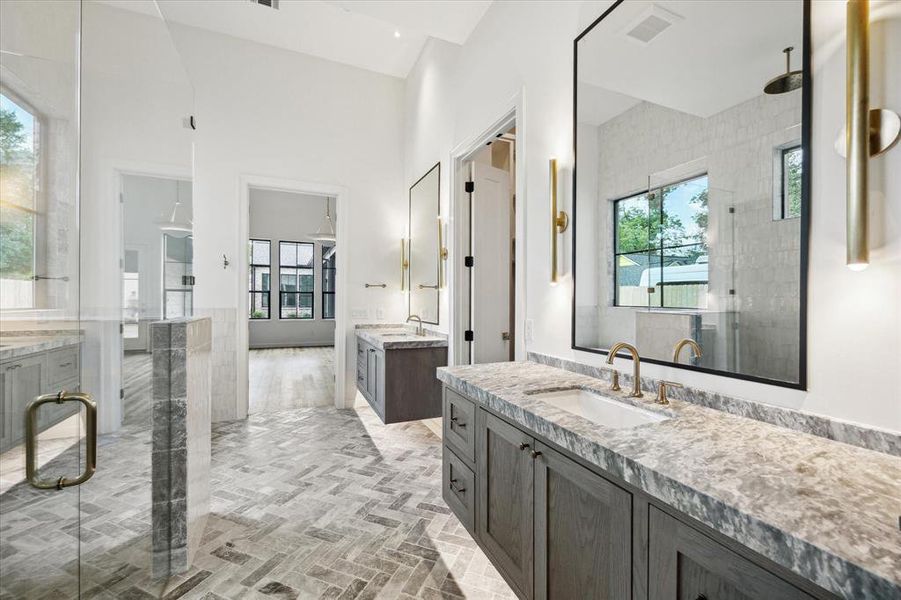 This elegant bathroom showcases floating vanities with marble countertops, accompanied by sleek brass fixtures. The glass-enclosed shower features a dual-showerhead setup, adding a touch of luxury. Large mirrors and modern sconces enhance the bright, airy feel of the space, while the herringbone tile flooring adds a stylish, cohesive element. The bathroom seamlessly connects to the adjacent bedroom, and two walk-in closets.